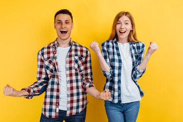 Obraz premium young man and woman, in checkered shirts and white t-shirts, celebrate the victory, showing a victory gesture, on a yellow background