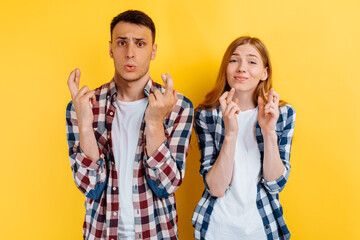 Intrigued young couple, man and woman praying with crossed fingers on yellow background