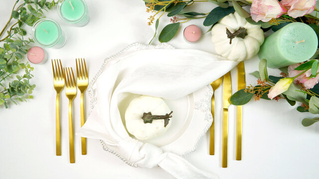 Happy Thanksgiving table setting with white vintage style plates, gold silverware, and centerpiece with greenery, pink, and white pumpkins, on white tablecloth. Top down view overhead styled flat lay.