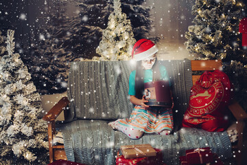 Portrait of lovely girl in Santa s red hat opening box with magical light inside sitting over blanket on couch surrounded by Christmas trees in snow..
