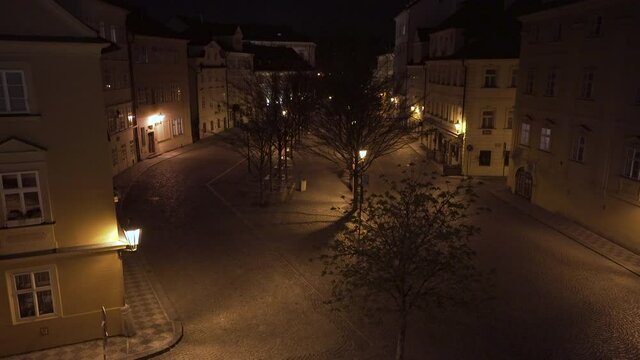 Empty city square Kampa in Prague, Czechia, trees and deserted streets during a Covid-19 lockdown at night, houses and pavement lit by street lanterns, romantic atmosphere, shot through iron railing.