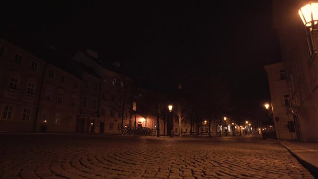 Empty city square Kampa of paving stones in Prague, Czechia, with trees, old houses and street lanterns shining above during a Covid-19 lockdown at night, illuminated by streetlights, slow pan shot.