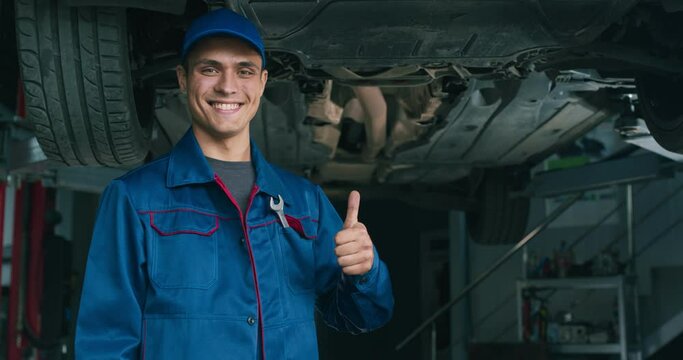 Friendly positive auto service worker smiling to camera and showing thumb up gesture, approving repair workshop