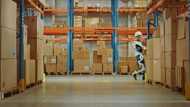 High-Tech Futuristic Warehouse: Manager Scans Packages for Inventory, Delivery in the Background Worker Wearing Advanced Full Body Powered exoskeleton, Walks with Heavy Pallet full of Cardboard Boxes