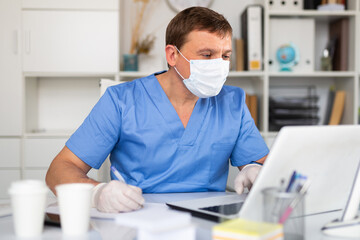 Fototapeta na wymiar Confident male doctor in blue medical uniform, mask and gloves working on laptop in clinic office