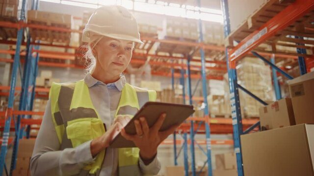 Professional Confident Worker Wearing Hard Hat Checks Stock and Inventory with Digital Tablet Computer in the Retail Warehouse full of Shelves with Goods. Working in Logistics, Distribution Center