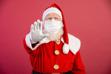 Fototapeta na wymiar Real Santa Claus on red background, wearing protective mask and with an open hand in front making a pandemic stop sign. Christmas with social distance. Covid-19