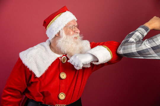 Real Santa Claus with red background, wearing glasses and hat doing elbow salute. Christmas with social distance. Covid-19