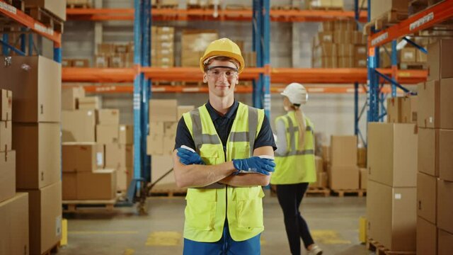 Handsome Smiling Worker Wearing Hard Hat, Crossing Arms Standing in the Retail Warehouse full of Shelves with Cardboard Boxes. Professional Working in Logistics, Delivery, and Distribution Center