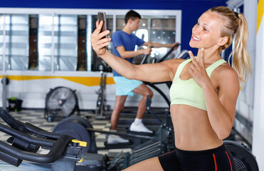 Naklejka premium Positive smiling friendly sporty girl taking selfie during workout at gym