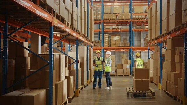 Retail Warehouse full of Shelves with Goods in Cardboard Boxes, Male and Female Managers Use Digital Tablet, Talk about Product Delivery. In Background Forklift Working in Logistics Storehouse