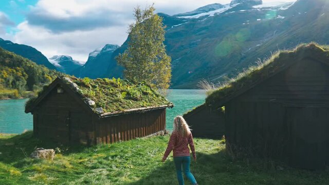 Camera slightly panning and following a girl walking through an abandoned old village, watching over a scenic lake and mountain view in Loen, Norway.