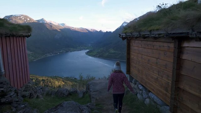 amera following a girl walking past a small cabin, towards beautiful view point, watching over a big lake with mountains in Loen, Norway.