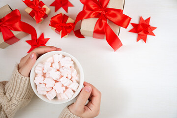 Female hands hold a cup of coffee with marshmallows and gifts with a red bow on a white background. Christmas and feminine desktop concept. Flat lay, top view.