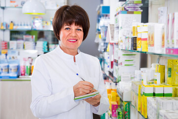 Fototapeta premium Portrait of cheerful smiling female druggist in white coat working in pharmacy