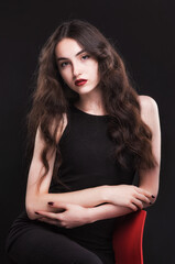 Fototapeta premium Beautiful slender young brunette with bright makeup in a black dress on a red chair