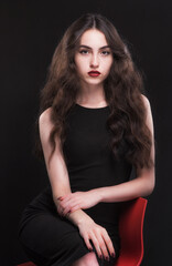Naklejka premium Beautiful slender young brunette with bright makeup in a black dress on a red chair