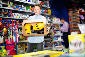 Serious happy cheerful smiling teen boy looking narrowly at yellow plastic car in children toy store