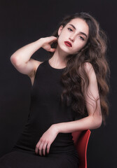 Naklejka premium Beautiful slender young brunette with bright makeup in a black dress on a red chair