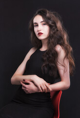 Naklejka premium Beautiful slender young brunette with bright makeup in a black dress on a red chair