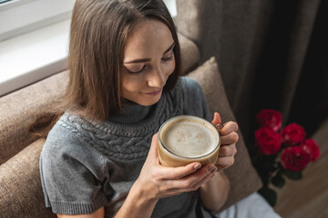 A young beautiful girl is sitting on the bed by the window and enjoying coffee in a cozy...