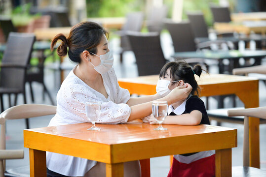 Mother fixing mask on child in outdoor restaurant.Young female and kid with facemask. Social distancing in public.People waiting to be served on the table in bar setting.Pandemic Covid19.