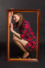 Studio photo of a young woman with long brown hair in a red checked shirt and bare legs. Squatting in a wooden picture frame