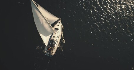 Top down of yacht under white sail at dark open sea aerial. Ocean bay black water and lonely sailboat. Summer cruise on boat. Luxury yachting scenery at serene seascape in dramatic drone shot