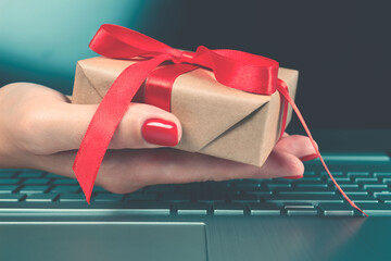 Woman's hand holding present box with red bow and laptoptop computer as background behind it for...