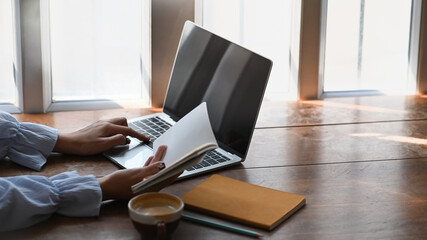 Side view of college student is reading book to find information and using laptop on wooden desk.