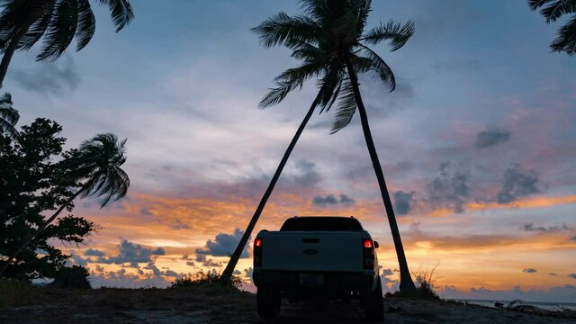 Silhouette of a pickup truck standing on the beach between crossed palm trees against the backdrop of a breathtaking sunrise. Flying colorful clouds in the sky. 4k time-lapse