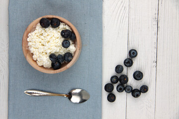 wooden bowl of farmer cheese with a spoon on blue napkin and a pile of blueberry on white wooden background flat lay. Healthy eating concept. Horizontal image