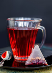 fruit tea pyramid bag and a transparent glass cup of red hibiscus drink, fruit pieces and rosehips in tea bag on dark background