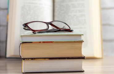 A stack of books with glasses and an open book in the background.