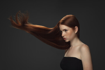 Beautiful model with long smooth, flying red hair isolated on dark grey studio background. Young...
