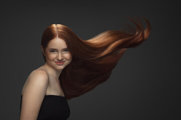 Beautiful model with long smooth, flying red hair isolated on dark grey studio background. Young caucasian girl with well-kept skin and hair blowing on air. Concept of salon care, beauty, fashion.