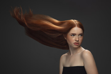 Beautiful model with long smooth, flying red hair isolated on dark grey studio background. Young caucasian girl with well-kept skin and hair blowing on air. Concept of salon care, beauty, fashion.