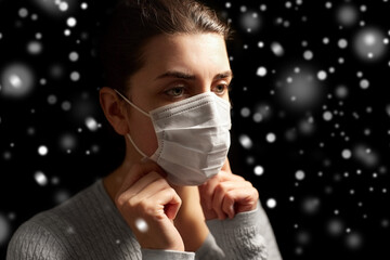 health protection, safety and pandemic concept - sick young woman adjusting protective medical face mask over snow on black background in winter
