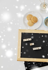 quarantine, epidemic and safety concept - close up of chalkboard with stay at home words on wooden toy blocks, coffee cup, cookies and book on white background in winter over snow