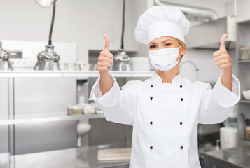 Fototapeta premium cooking, culinary and health concept - female chef in toque wearing face protective medical mask for protection from virus disease showing thumbs up over restaurant kitchen background
