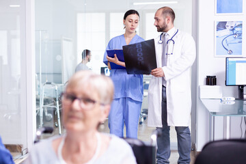 Obraz premium Doctor holding patient x-ray explaining diagnosis to nurse in waiting area. Disabled senior woman wheelchair waiting for medical examination. Hospital and health care system, medicine private clinic