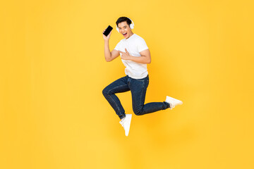 Fototapeta na wymiar Happy young Asian man wearing headphones listening to music from mobile phone and jumping with hand poiting isolated on yellow background