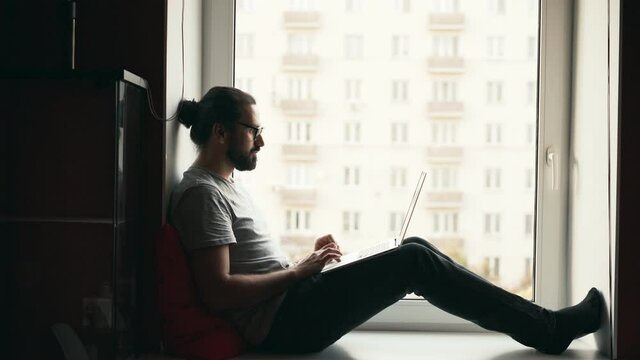 Handheld shot of a young handsome man working on his laptop while sitting on a windowsill with a city view. Freelancer working from home.