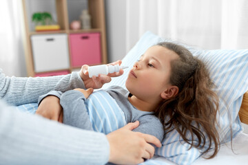 Obraz na płótnie Canvas family, health and medicine concept - mother with nasal spray treats little sick daughter lying in bed at home