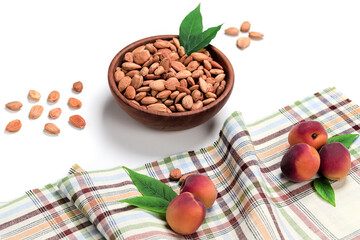 Peeled apricot kernels in a wooden bowl, fresh apricots with green leaves on checkered tablecloth isolated on white background. Including clipping path