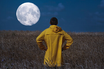 young man standing in a wheat field back to the camera in a night and watching super moon in sky