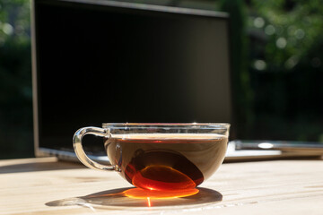 transparent cup of tea with shining ray of sun light in it, laptop and phone not in focus on light wooden table. Remote work concept. Working in cozy place