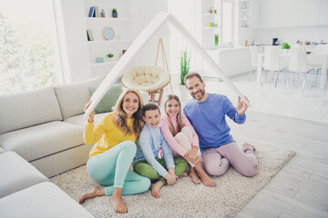 Full size photo of harmony family sit floor mom dad hold paper card roof above two little kids girl boy in house indoors