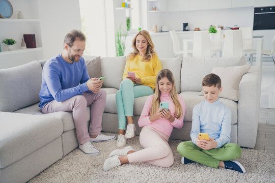 Full length photo of mommy daddy and two little kids texting on smartphone sit sofa floor in house indoors