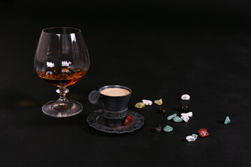 A glass of cognac, brandy or whiskey, little cup of coffee of dark metal and a chocolate sweets, scattered on black background. Copy space.